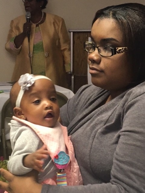 Mother holding baby at ministry event with speaker in background