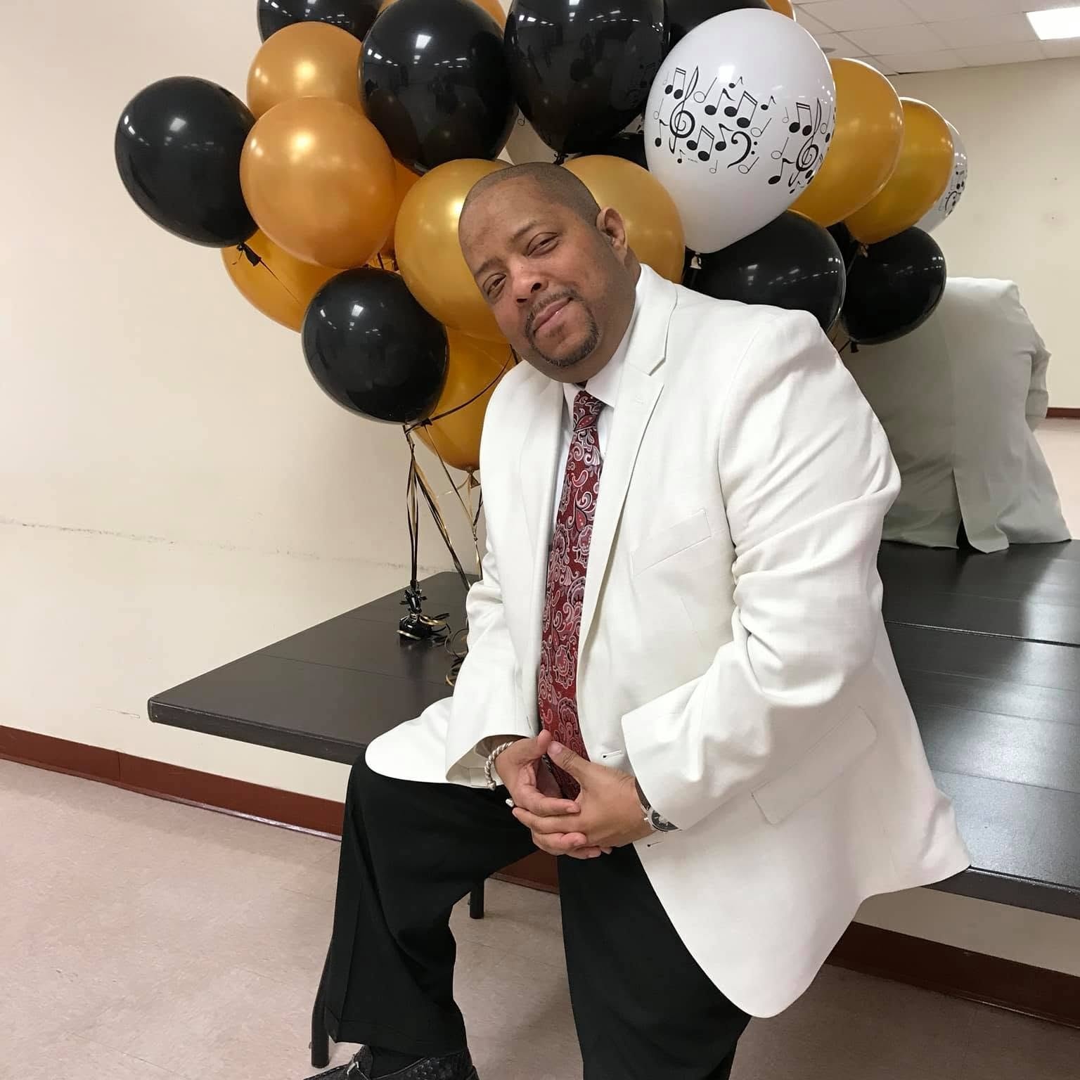 John Tillery in white jacket seated in front of gold and black balloons